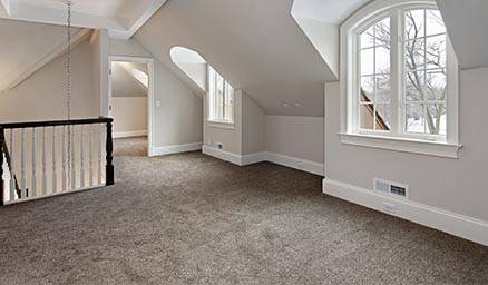 Empty attic room with carpet flooring, white walls, dormer windows, and a railing overlooking stairs